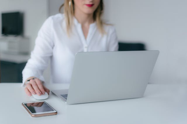 A woman using a silver laptop beside an iPhone.