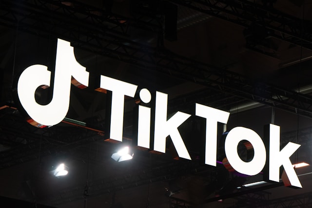 An illuminated TikTok logo sign hangs from the ceiling in a dark indoor setting, with overhead lights.