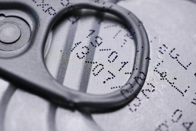 A close-up view of a beverage can's pull tab, with expiry date and dotted printed codes visible on the metallic surface.